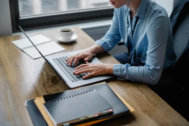 homem em uma mesa de escritório trabalhando em um notebook, com pastas, diário e uma chicara de café